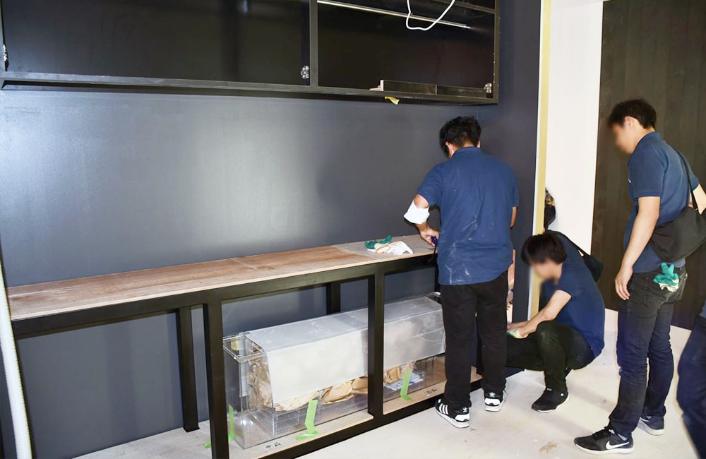 黒い壁面の前に大型水槽台を設置する作業員たち