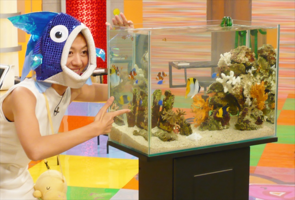 魚の被り物をした女性と海水魚が泳ぐ水槽