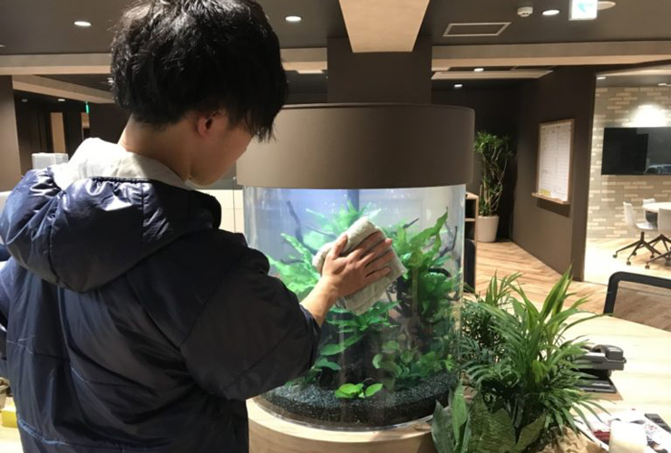 円柱水槽の表面を布で拭き掃除するスタッフの背中